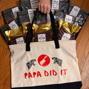 Hand holding a Papa Did It tote bag filled with basturma, pork loin, turkey balyk, and beef sticks
