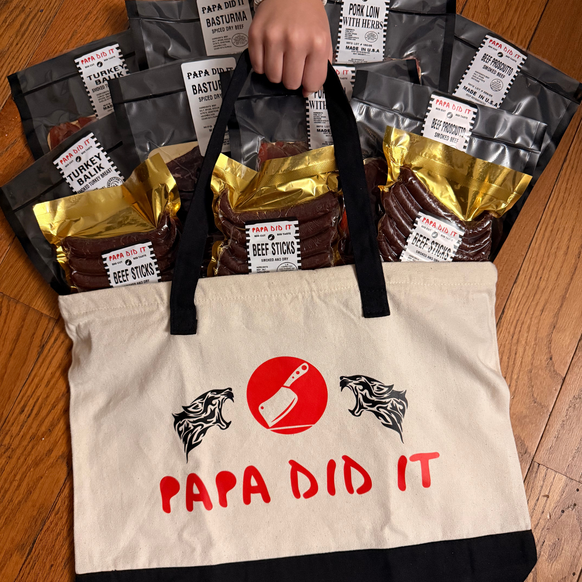 Hand holding a Papa Did It tote bag filled with basturma, pork loin, turkey balyk, and beef sticks