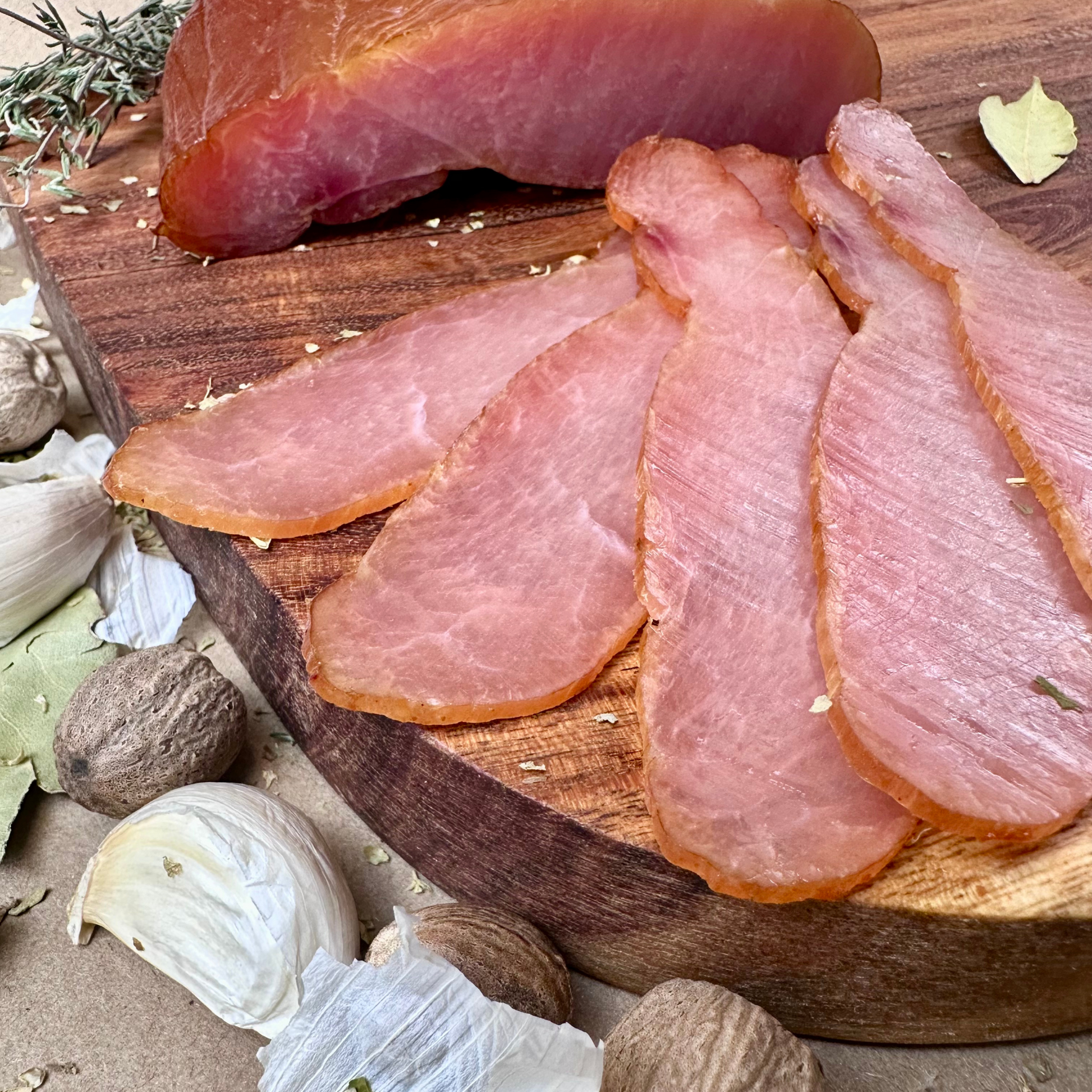 Sliced ham on a wooden cutting board with herbs and garlic cloves.