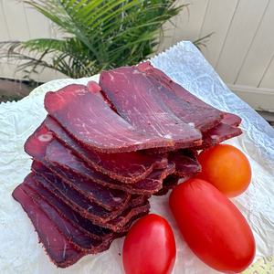 Stack of sliced cured meat with cherry tomatoes on a white surface outdoors.