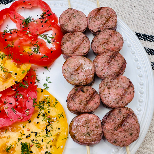 Grilled sausage skewers served with colorful heirloom tomatoes, herbs, and sesame seeds on a white plate