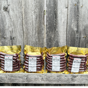 papa did it beef sticks lineup on wooden background — four golden vacuum-sealed packs of handcrafted hickory-smoked beef sticks made in the USA, displayed neatly against rustic wood for a clean artisan presentation