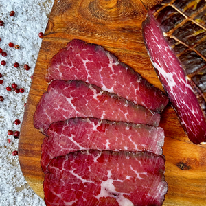 Detail shot of hickory-smoked beef prosciutto slices on a wooden board with sea salt and pink peppercorns — handcrafted dry-cured beef by Papa Did It.
