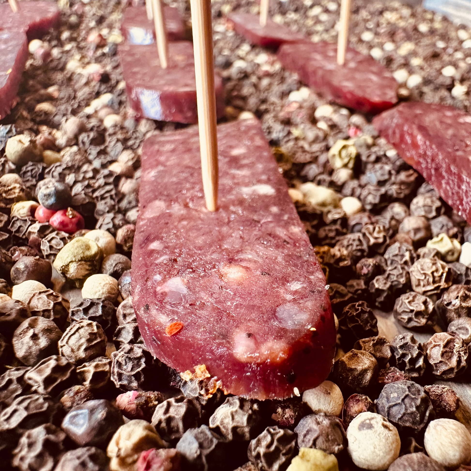 Close-up of sliced beef sujuk dry-cured sausage on skewer over peppercorns – artisanal hickory smoked beef slice