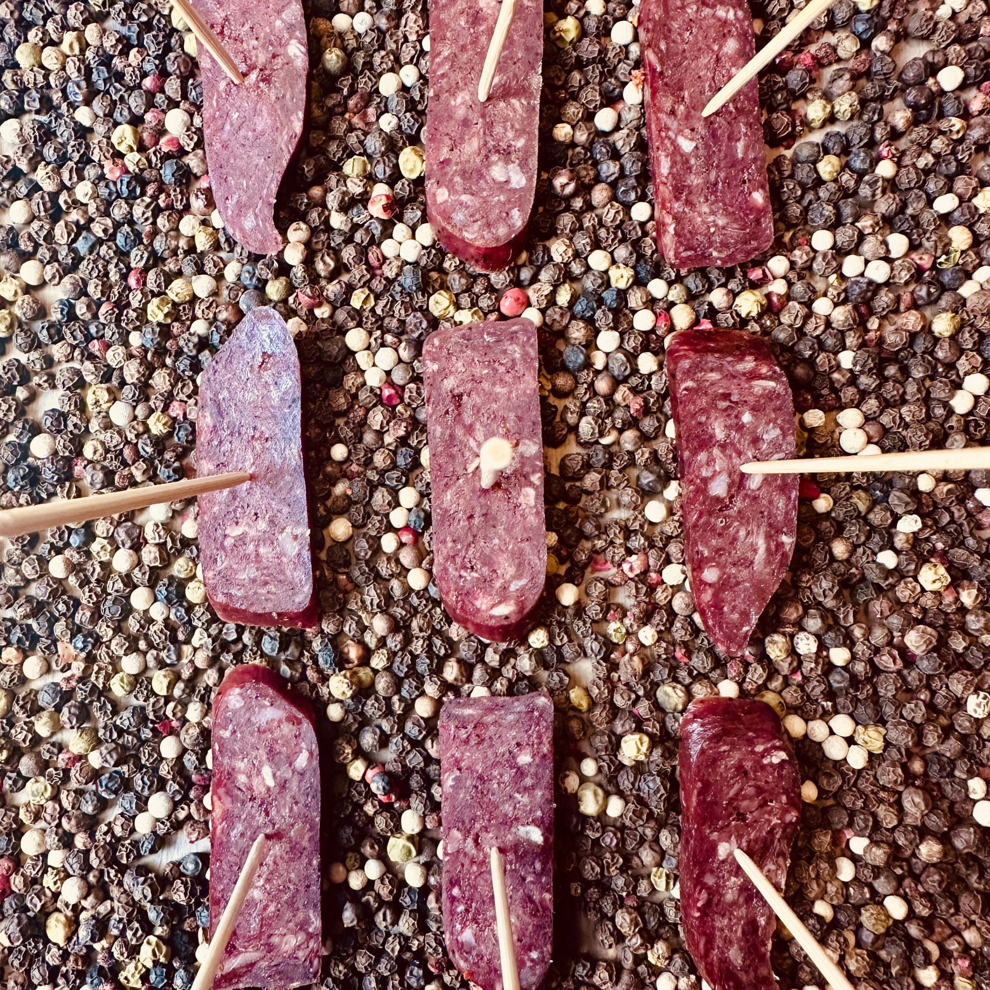 Top view of sliced beef sujuk dry-cured sausage on skewers over peppercorns – artisanal hickory smoked beef slices