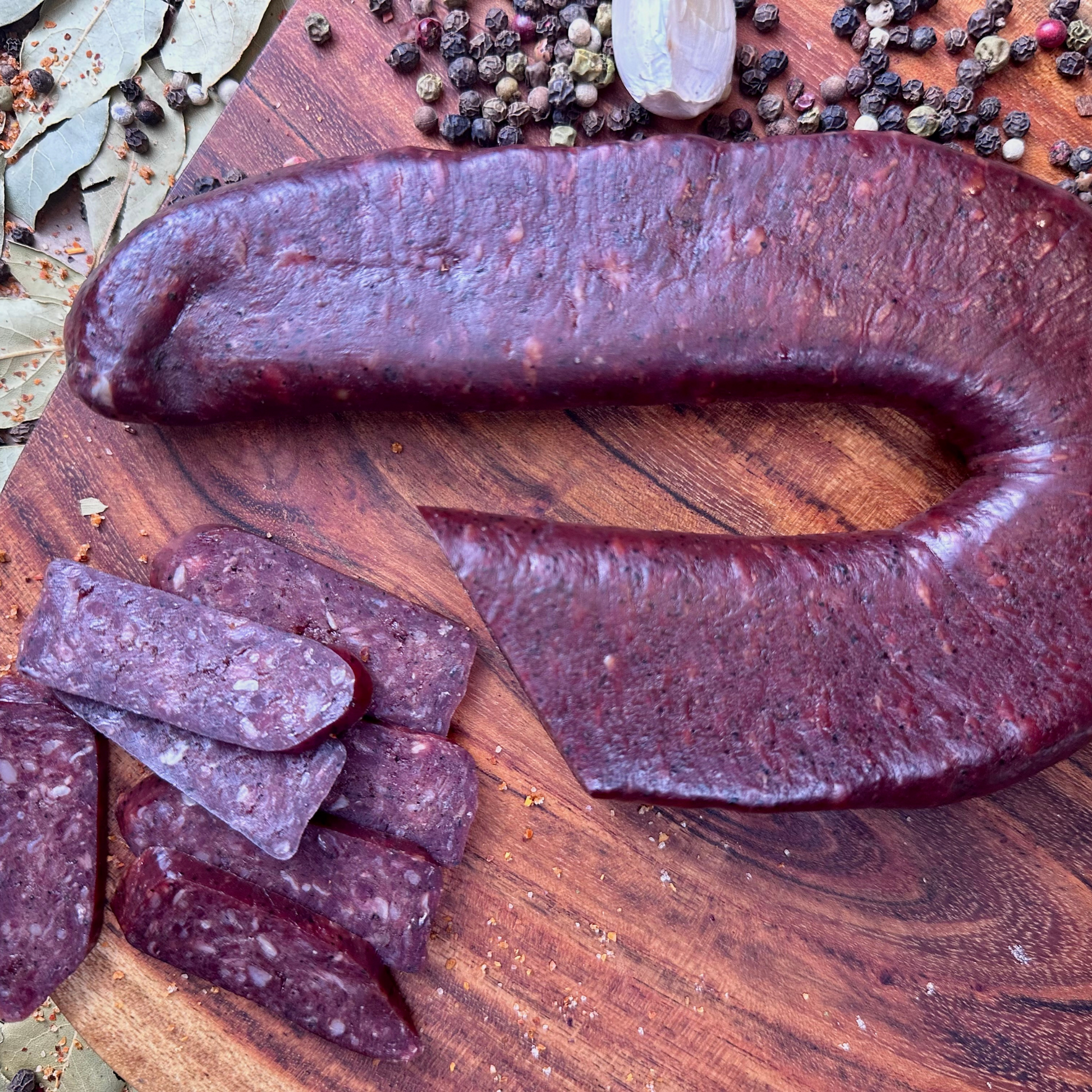 Whole and sliced beef sujuk dry-cured sausage on wooden board with peppercorns and spices – artisanal hickory smoked beef