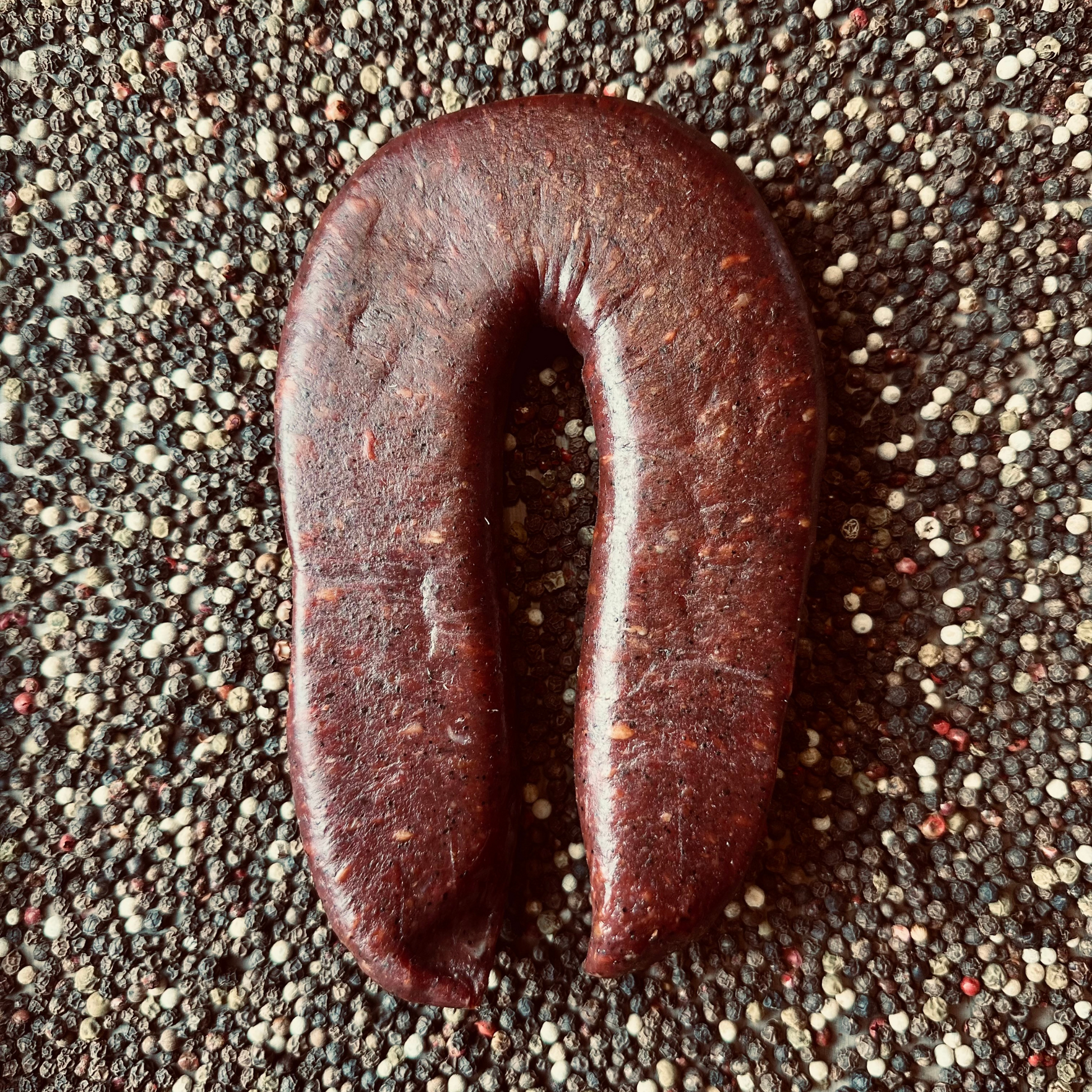 Whole beef sujuk dry-cured sausage in horseshoe shape on peppercorn background – artisanal hickory smoked sausage